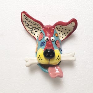 May include: A colorful ceramic dog head with a bone in its mouth. The dog has large ears, a black nose, and a pink tongue. The dog's head is painted with red, blue, yellow, and white accents.