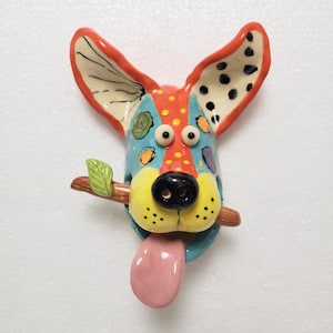 May include: A whimsical ceramic dog figurine with a colorful design. The dog has large, expressive eyes, a black nose, and a pink tongue sticking out. It holds a small branch with a green leaf in its mouth. The ears are red with black spots.