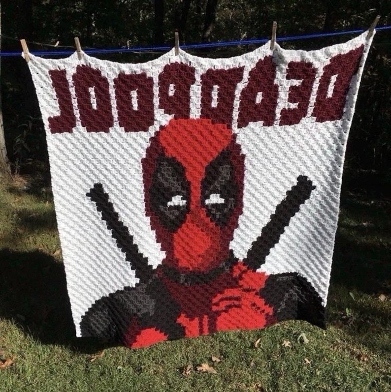May include: A white and red crocheted blanket featuring the character Deadpool. The word "DEADPOOL" is displayed in red block letters at the top. The blanket is hanging outside on a clothesline.