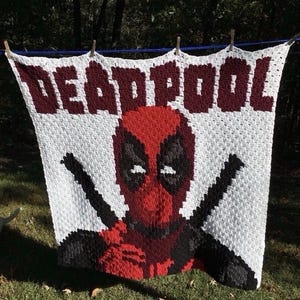 May include: A white crocheted blanket featuring the word "DEADPOOL" in maroon letters. The blanket displays a pixelated image of Deadpool's face in red, black, and white, with black crossed swords.