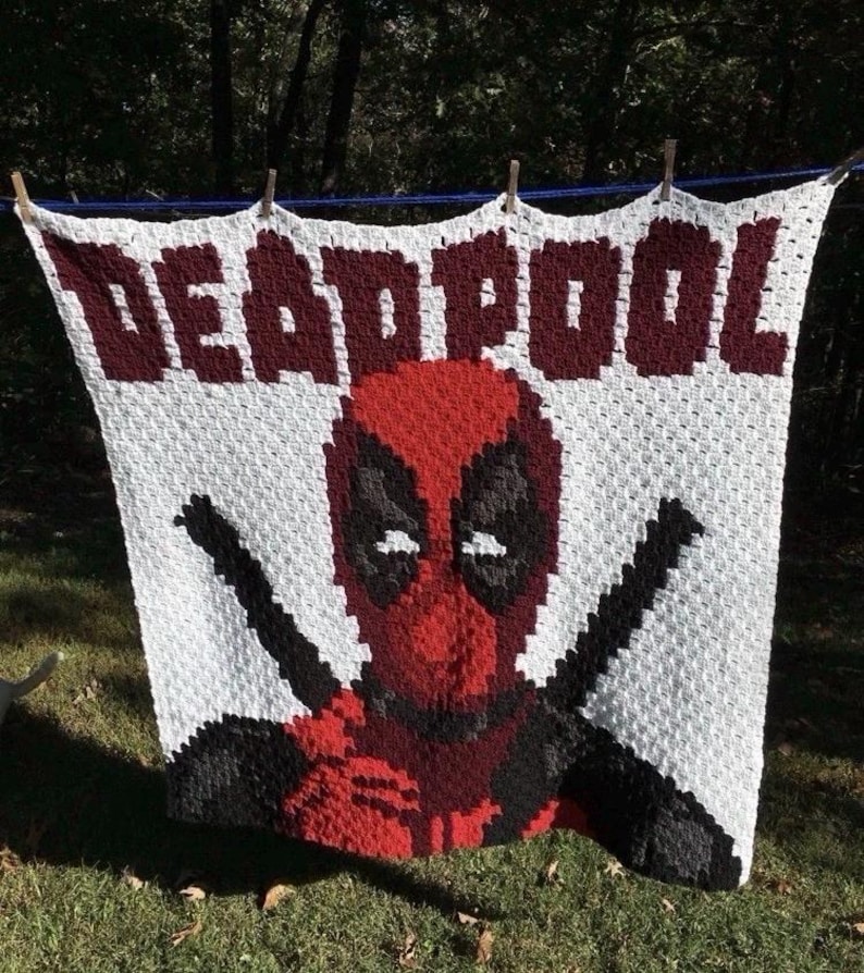 May include: A white crocheted blanket featuring the word "DEADPOOL" in maroon letters. The blanket displays a pixelated image of Deadpool's face in red, black, and white, with black crossed swords.