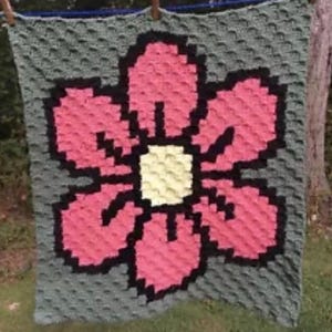 May include: A handmade crochet blanket featuring a large pink flower with a black outline and a yellow center. The flower is set against a gray background. The blanket is hanging outside on a clothesline.