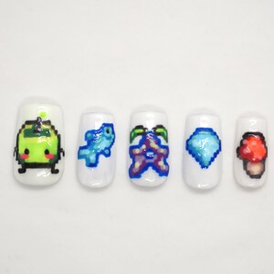 May include: Five white nail tips with different pixelated designs. The designs include a green creature, a blue fish, a purple starfish, a blue diamond, and a red mushroom.