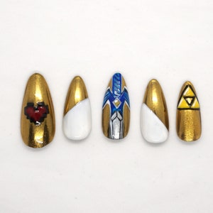 Puede incluir: Cinco uñas postizas de color dorado con diferentes diseños. Una uña tiene un corazón rojo pixelado, otra tiene un diseño blanco y dorado, otra tiene un diseño de espada azul y plata, otra tiene un diseño blanco y dorado y otra tiene un triángulo amarillo con un triángulo más pequeño dentro.