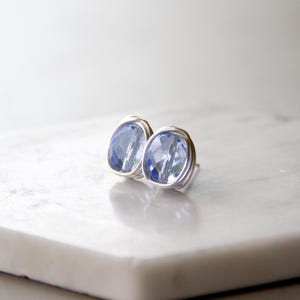 May include: A pair of stud earrings featuring faceted, light blue, cushion-cut crystals. Each crystal is securely encased in a delicate, coiled silver-toned wire frame that wraps around the perimeter of the stone, creating a textured, metallic border.