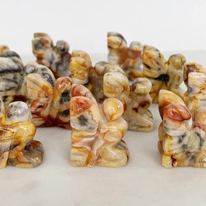 Könnte beinhalten: Eine Sammlung kleiner, geschnitzter Stein-Schmetterlingsfiguren. Die Schmetterlinge sind in verschiedenen Schattierungen von Beige, Creme, Grau und Rot gehalten und weisen komplizierte Muster auf. Die Figuren werden auf einer weißen Oberfläche präsentiert, um die natürlichen Farben und Texturen des Steins hervorzuheben.