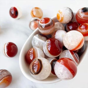 May include: A collection of polished agate gemstone spheres in a white bowl. The spheres display a variety of colors, including shades of red, brown, white, and orange. The gemstones have a banded appearance, with some showing translucent qualities.