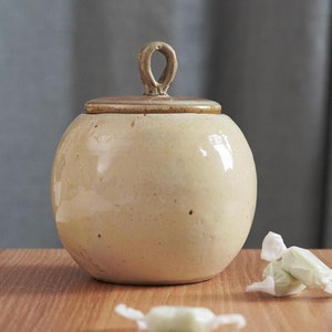 Puede incluir: Un tarro de cerámica con tapa y asa de bucle. El tarro es de color amarillo pálido con una tapa marrón. El tarro está sentado en una mesa de madera con varios caramelos verdes envueltos en papel blanco.