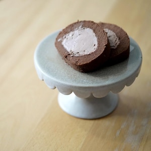 Könnte beinhalten: Zwei Stücke Schokoladenrollenkuchen mit einer hellbraunen Füllung werden auf einem weißen Keramik-Kuchenständer mit gewellten Rändern präsentiert. Der Kuchenständer steht auf einer Holzoberfläche.