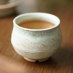 May include: A small, handcrafted ceramic cup with a textured, off-white exterior and a light green band near the rim. The cup contains a light brown liquid, possibly tea or coffee. The cup sits on a wooden surface.