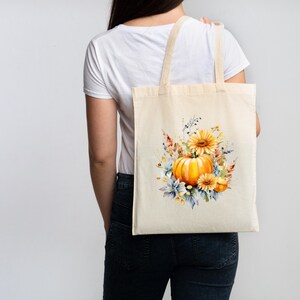 Pode incluir: Uma bolsa tote bege com um design de aquarela de uma abóbora, girassóis e outras folhas de outono.