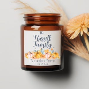 May include: A brown glass jar candle with a white label. The label has a watercolor design of pumpkins and fall leaves. The text on the label reads "The Russell Family Pumpkin Farm est. 2024".