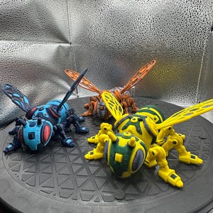 Op de afbeelding: Drie 3D-geprinte roboterbij-figuurtjes. Eén is blauw en zwart, één is geel en groen en één is oranje en zwart.