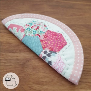 May include: A half-circle quilted coaster with a pink and white polka dot border. The coaster is made of fabric scraps in various floral patterns, including pink, green, blue, and white.