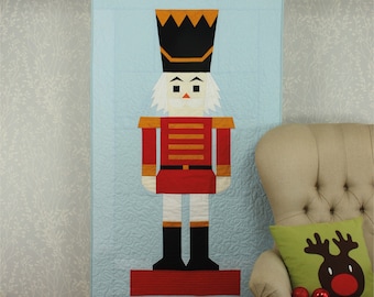 May include: A blue quilted wall hanging featuring a red and white nutcracker with a black hat and gold accents. The nutcracker is standing on a red platform.