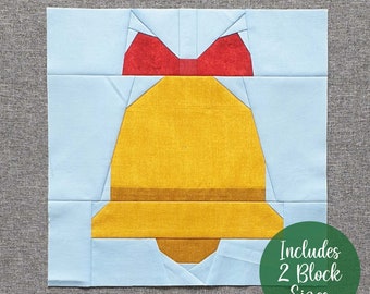 Op de afbeelding: Een papieren patchwork quiltblokpatroon met een gele bel met een rode strik erop. Het patroon omvat twee blokformaten. De tekst "Includes 2 Block Sizes" is gedrukt op een groene cirkel.