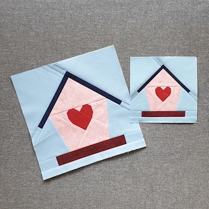 May include: Two paper pieced quilt blocks in the shape of houses with red hearts. The larger block is light blue with a pink house and a burgundy base. The smaller block is light blue with a pink house and a burgundy base.
