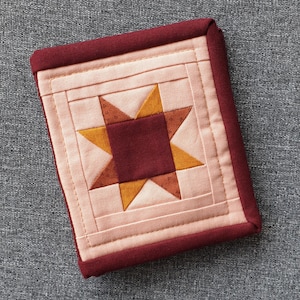 May include: A pink and red quilted fabric coaster with a star design in the center. The star is made of burgundy, orange, and pink fabric.