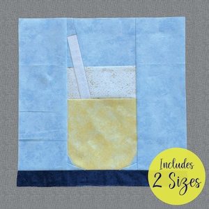 Puede incluir: Un bloque de colcha de patchwork azul y amarillo con un vaso de limonada con una pajita. El bloque está rodeado por un borde azul y un borde inferior azul oscuro. El texto "Includes 2 Sizes" está en un círculo amarillo en la esquina inferior derecha.