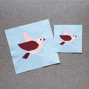 May include: Two paper pieced bird quilt blocks. The larger block is light blue with a pink and maroon bird. The smaller block is light blue with a pink and maroon bird.