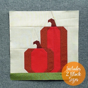 Puede incluir: Un bloque de tela con dos calabazas estilizadas en tonos rojos y marrones, con verde en la base. Las calabazas están sobre un fondo crema. El texto "Includes 2 Block Sizes" está en un círculo naranja.
