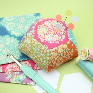 Peut inclure: Un coussin à épingles en patchwork coloré de forme hexagonale, rempli d'épingles avec des têtes en plastique colorées. Le coussin à épingles est posé sur une surface verte avec un morceau de tissu, un découseur et une bobine de fil.