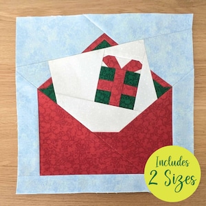 May include: A fabric Christmas-themed block featuring a red envelope with a white card and a green and red gift. The block is bordered by light blue fabric and includes the text "Includes 2 Sizes" in a yellow circle.