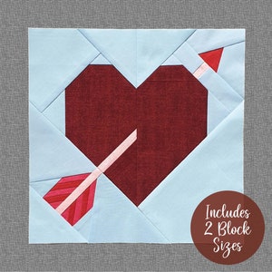 Op de afbeelding: Een quiltblokpatroon met een rood hart met een roze en rode pijl erdoorheen. De achtergrond is lichtblauw. De tekst "Includes 2 Block Sizes" staat in een bruine cirkel.