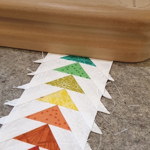 May include: A row of colorful fabric triangles sewn together in a pattern. The triangles are arranged in a rainbow order, with pink at the bottom and green at the top. The fabric is white with a floral pattern.