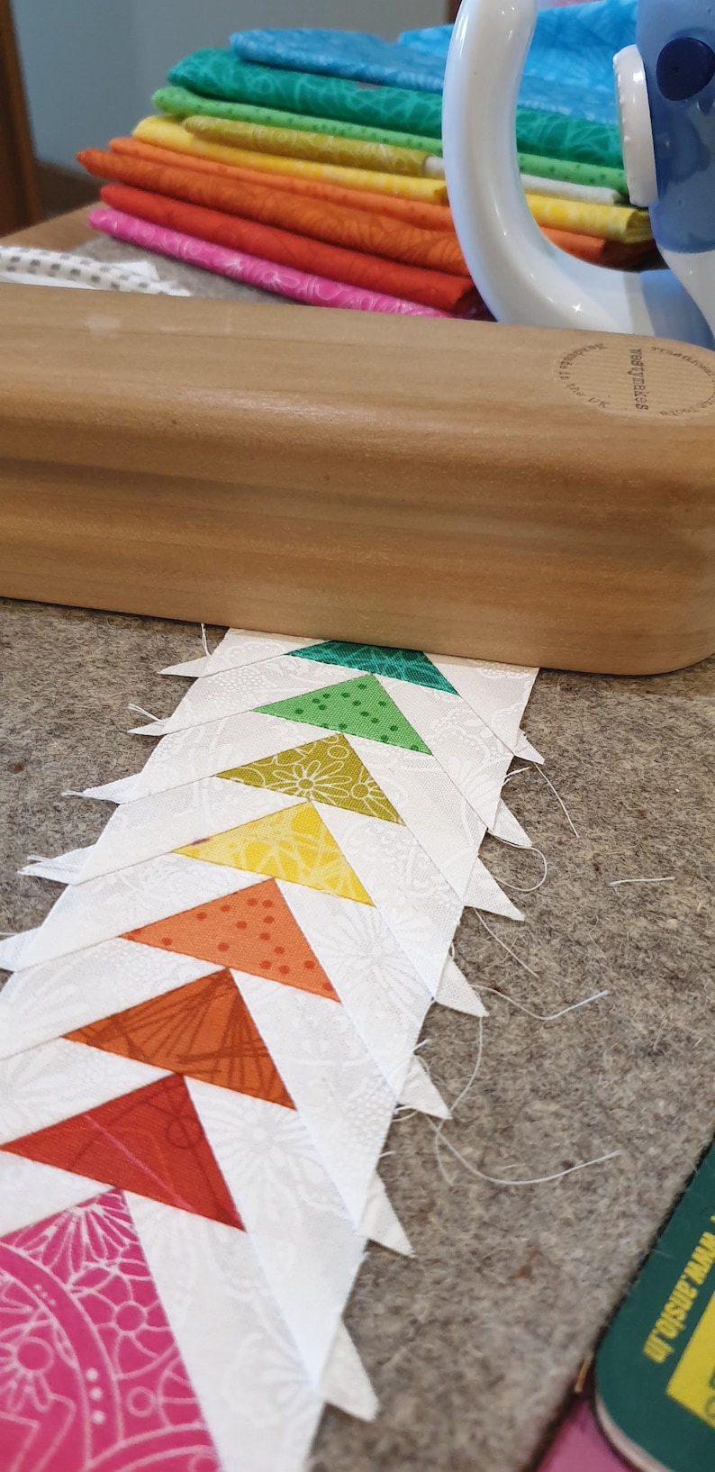 May include: A row of colorful fabric triangles sewn together in a pattern. The triangles are arranged in a rainbow order, with pink at the bottom and green at the top. The fabric is white with a floral pattern.