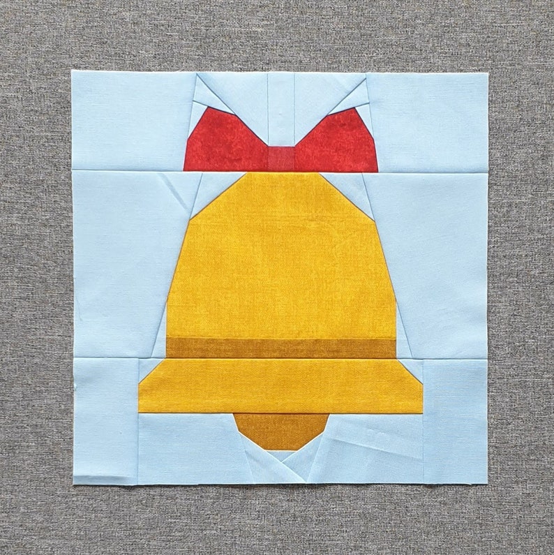 Op de afbeelding: Een gele bel met een rode strik erboven. De bel is gemaakt van stof en maakt deel uit van een patchwork quiltblok.