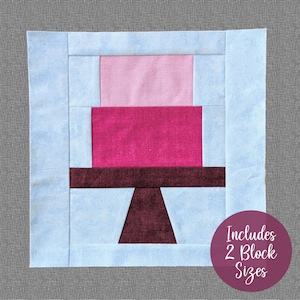 May include: A quilt block pattern featuring a cake design with pink, red, and maroon fabric. The block is set against a light blue background. The text "Includes 2 Block Sizes" is visible in the lower right corner.