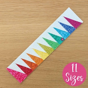 Op de afbeelding: Een kleurrijke patchwork strook met afwisselend witte en regenboogkleurige driehoeken in een zigzagpatroon. Een roze cirkel met de tekst "11 Sizes" is zichtbaar. De strook ligt op een houten oppervlak. De driehoeken zijn van stof.