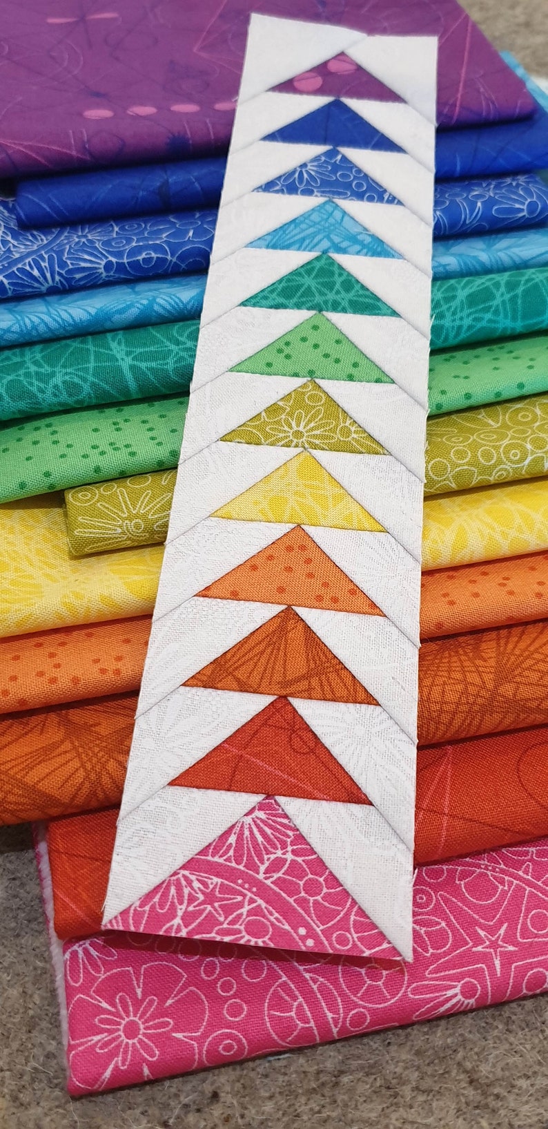 May include: A white fabric strip with a row of colorful fabric triangles sewn on top. The triangles are in a rainbow pattern, starting with purple and ending with pink.