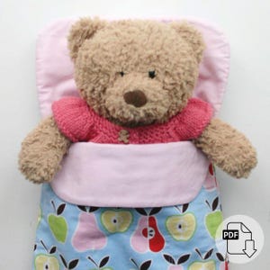 May include: A brown teddy bear wearing a pink sweater is tucked into a blue and pink sleeping bag with a pattern of apples and pears. The sleeping bag has a pink lining.