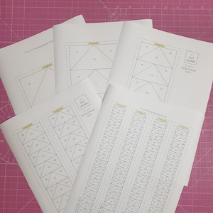 May include: Four sheets of paper with black lines outlining different sizes of flying geese quilt blocks. The sheets are labeled with the size of the finished square and the dimensions of the flying geese units. The sheets are on a pink cutting mat.