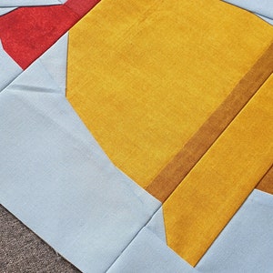 Op de afbeelding: Een quiltblok met een geel, bruin en rood ontwerp. Het blok is gemaakt van stof en heeft een strik in de linkerbovenhoek.
