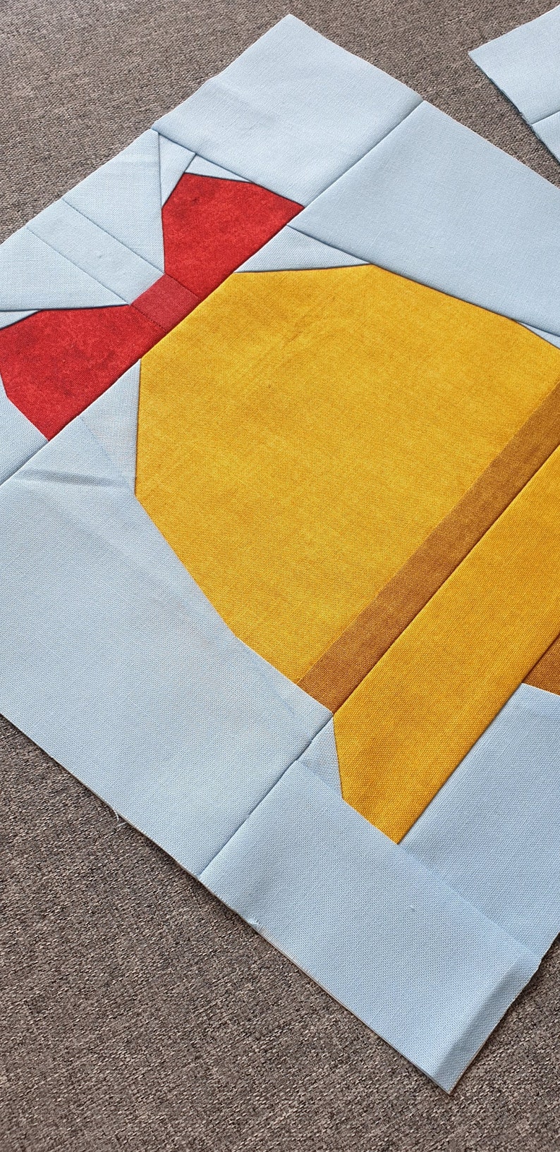 Op de afbeelding: Een quiltblok met een geel, bruin en rood ontwerp. Het blok is gemaakt van stof en heeft een strik in de linkerbovenhoek.