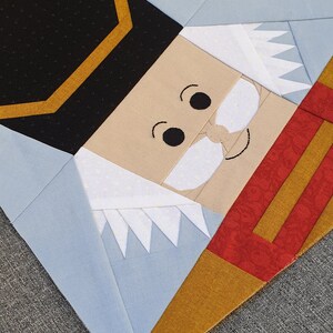May include: A patchwork quilt block featuring a nutcracker face. The face is made of beige fabric with black eyes, a white mustache, and a white beard. The nutcracker is wearing a black hat with a gold trim and a red and gold uniform.