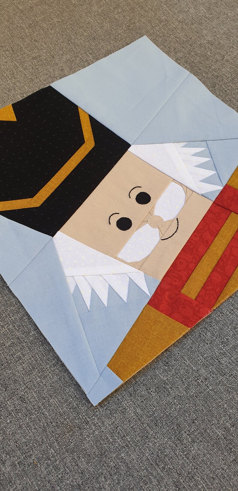 May include: A patchwork quilt block featuring a nutcracker face. The face is made of beige fabric with black eyes, a white mustache, and a white beard. The nutcracker is wearing a black hat with a gold trim and a red and gold uniform.