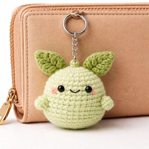 May include: A crocheted keychain in the shape of a smiling, light green fruit with two green leaves. The keychain has black eyes and rosy cheeks. It is attached to a silver keyring and chain. The background is a light pink wallet.