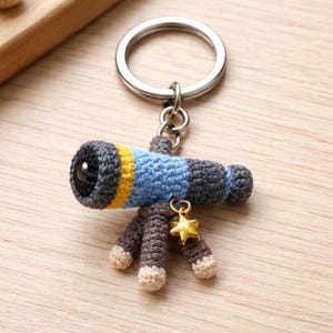 May include: Crocheted telescope keychain with a silver keyring. The telescope is gray, blue, and yellow, with a gold star charm. The tripod legs are brown with cream-colored feet. A unique accessory for astronomy enthusiasts.