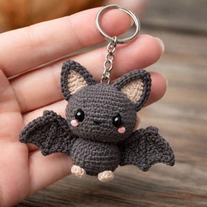 Peut inclure: Un porte-clés en forme de chauve-souris crochetée, de couleur gris foncé avec des détails beiges. La chauve-souris a de grands yeux noirs, des joues roses et de petits pieds beiges. Les ailes sont déployées, et le porte-clés a une chaîne et un anneau argentés.