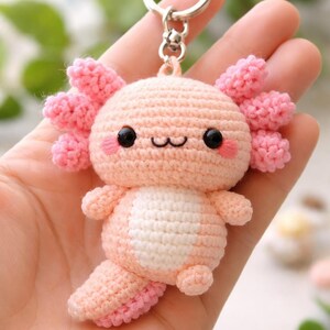 Op de afbeelding: Een handgemaakte gehaakte axolotl-sleutelhanger in roze en wit. De axolotl heeft zwarte ogen, roze wangen en een lachende mond. De sleutelhanger is bevestigd aan een zilverkleurige ring.