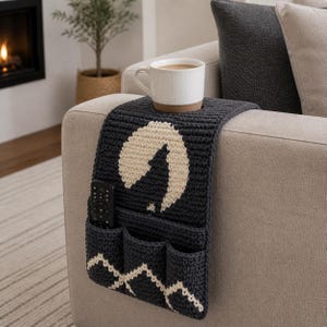 May include: A dark gray crocheted sofa armrest with a cream-colored moon design and three pockets. A white mug with coffee sits on top. A remote control is in one of the pockets. The armrest is on a beige sofa.