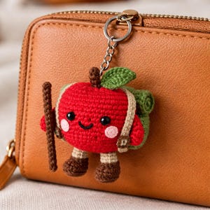 May include: A red crocheted apple keychain with a green leaf, brown stem, and a small backpack. The apple has a smiling face, pink cheeks, and brown boots. A brown walking stick is held in its hand.