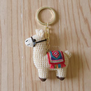 Könnte beinhalten: Ein gehäkeltes Lama-Schlüsselanhänger in Creme mit braunen Hufen und einer bunten Decke. Das Lama hat ein schwarzes Halfter und einen goldfarbenen Schlüsselring und eine Kette. Der Hintergrund ist eine hellbraune Holzoberfläche.