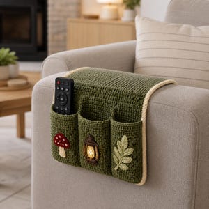 May include: A green crocheted sofa arm organizer with three pockets. The pockets are decorated with a mushroom, a lantern, and a leaf. A black remote control is in one of the pockets. The organizer has a cream-colored border.