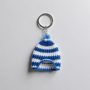 May include: A handmade crochet beanie keychain in blue and white stripes. The miniature hat features a blue pom-pom and is attached to a silver keyring. The keychain is a small, decorative accessory.