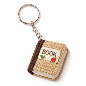 Puede incluir: Un llavero de libro tejido a ganchillo con un llavero plateado. El libro es de color beige con el lomo marrón y una etiqueta blanca con la palabra "BOOK" y una manzana y una hoja bordadas. Un accesorio encantador.